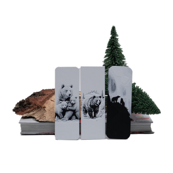 Three bookmarks depicting Bears sitting on books with wood and evergreen behind.