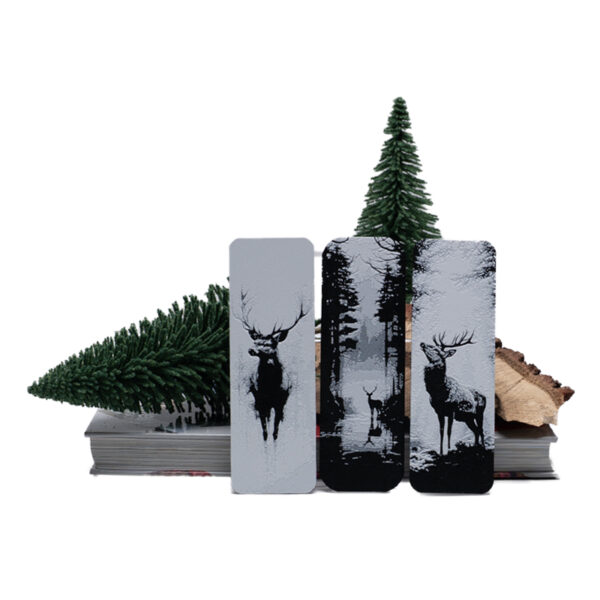 3 bookmarks wit bucks and stags in natural setting sitting on a stack of books with wood and evergreen trees behind them.